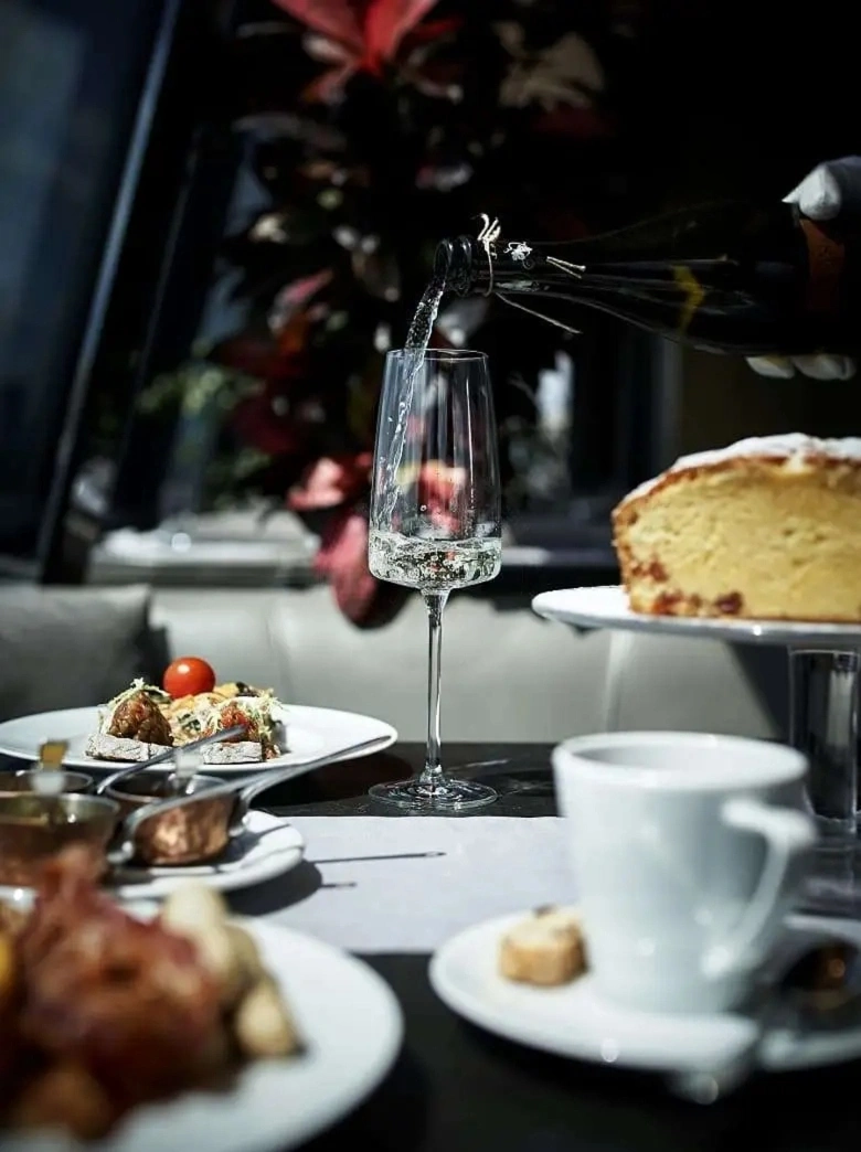 Un primer plano del servicio de un evento corporativo de Catering Vicente, que muestra a un camarero con guante blanco vertiendo vino espumoso en una flauta alta, con un bodegón de brunch de lujo de tartines, pasteles artesanos y café en el fondo.