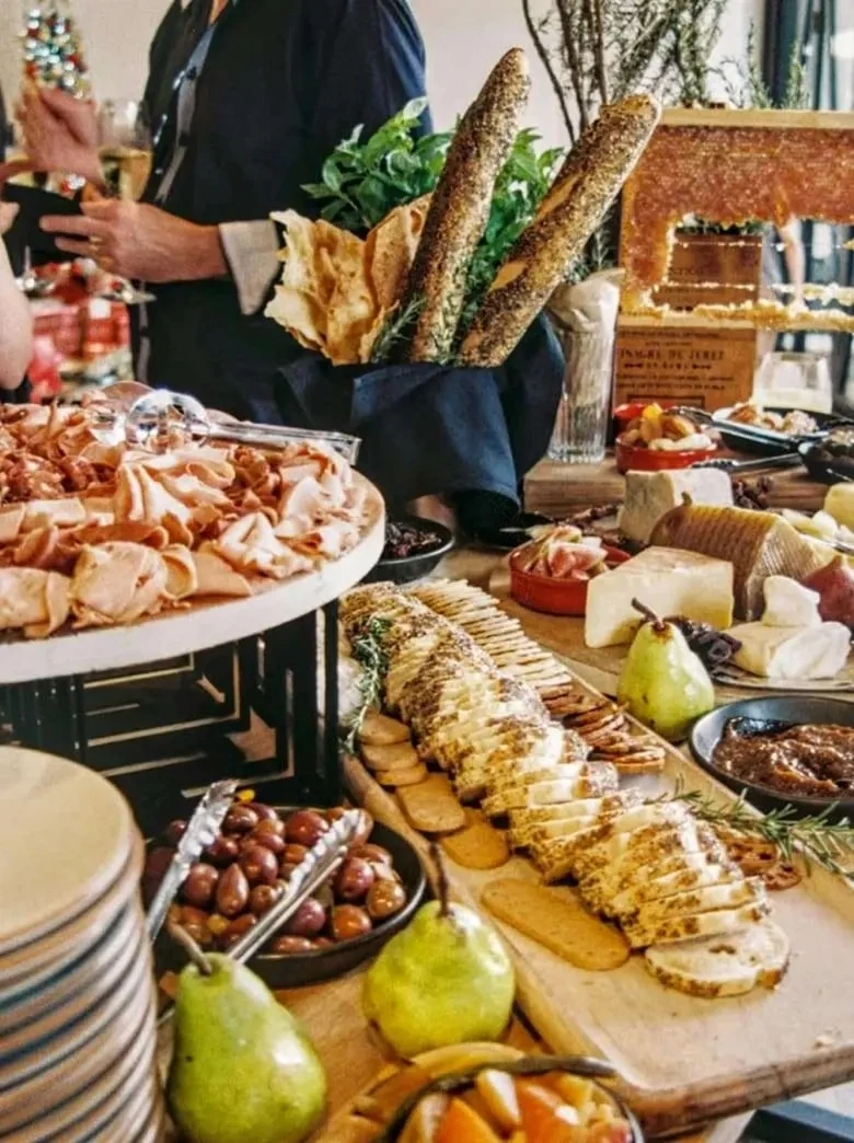 Un suntuoso buffet de catering de Catering Vicente en un evento festivo, que presenta un abundante y hermoso surtido de embutidos, quesos artesanales, panes artesanales, frutas frescas (como peras e higos) y aceitunas sazonadas.