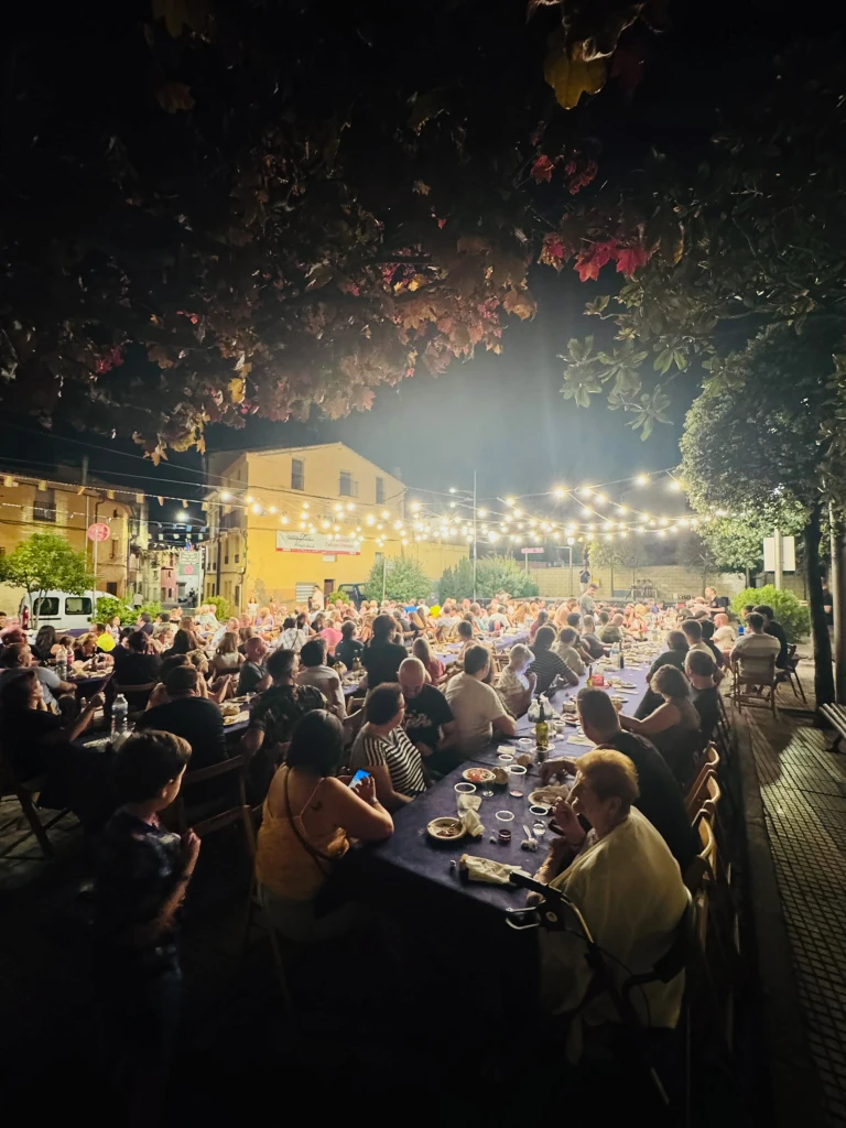 Fotografía nocturna de una gran cena popular al aire libre durante las fiestas de Tudelilla. Una multitud de personas está sentada a lo largo de extensas mesas con manteles azules, todo iluminado por guirnaldas de luces bajo las ramas de los árboles.