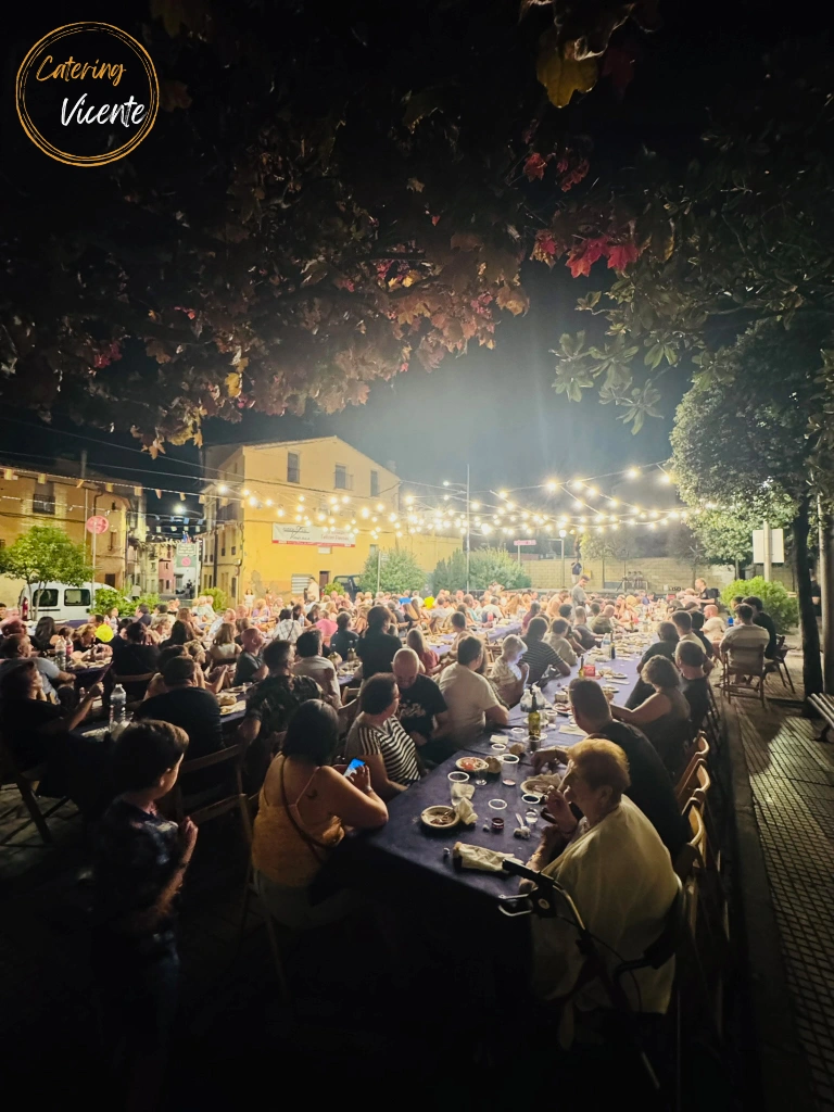 Fotografía nocturna de una gran cena popular al aire libre durante las fiestas de Tudelilla. Una multitud de personas está sentada a lo largo de extensas mesas con manteles azules, todo iluminado por guirnaldas de luces bajo las ramas de los árboles.