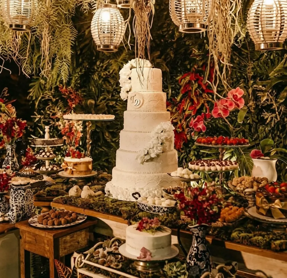 Una toma de media distancia de una elaborada y exuberante exhibición de postres de temática de jardín, creada por Catering Vicente para un momento único. El centro es un gran pastel de bodas blanco de siete pisos con encaje y rosas de azúcar, rodeado por innumerables postres en miniatura sobre mesas cubiertas de musgo, todo bajo el cálido resplandor de linternas de ratán colgantes y un exuberante muro de follaje.